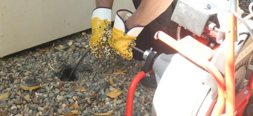 Beaver Brothers Plumbing drain machine feeding a cable into an outdoor cleanout in gravel beside a house