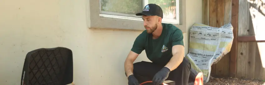 Beaver Brothers Plumbing technician in a branded green shirt and cap working beside a home's exterior wall